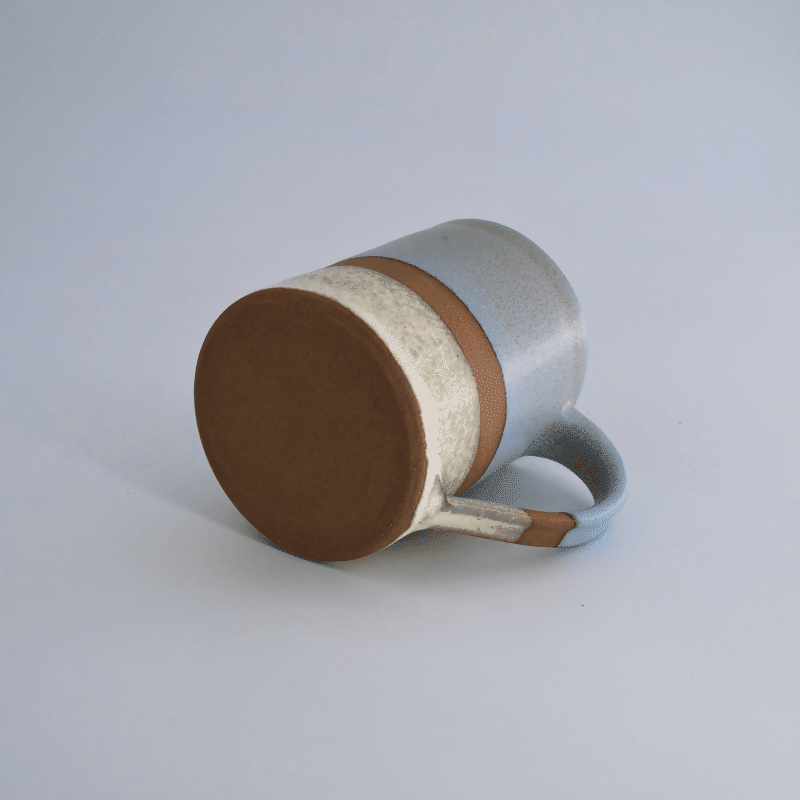Mug en grès rouge, émaillé bleu sur la partie supérieure et blanc sur la partie inférieure, avec une bande de séparation non émaillée laissant la terre apparente. Présenté couché sur fond clair.