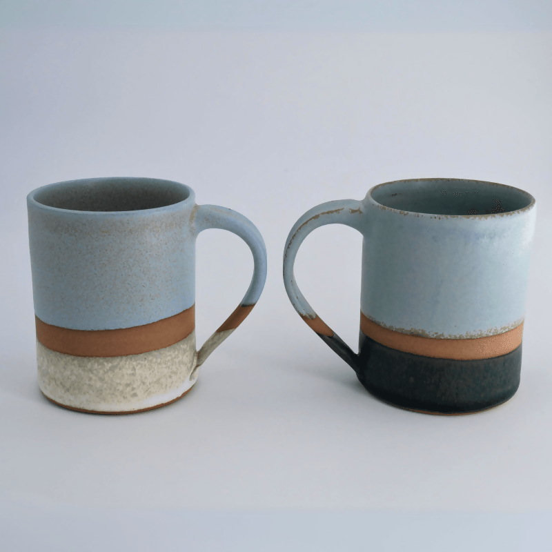 Duo de mugs en grès rouge, émaillé bleu et blanc pour celui de gauche, vert d'eau et noir pour celui de droite. chacun avec une bande de séparation non émaillée laissant la terre apparente. Présentés debouts sur fond clair.
