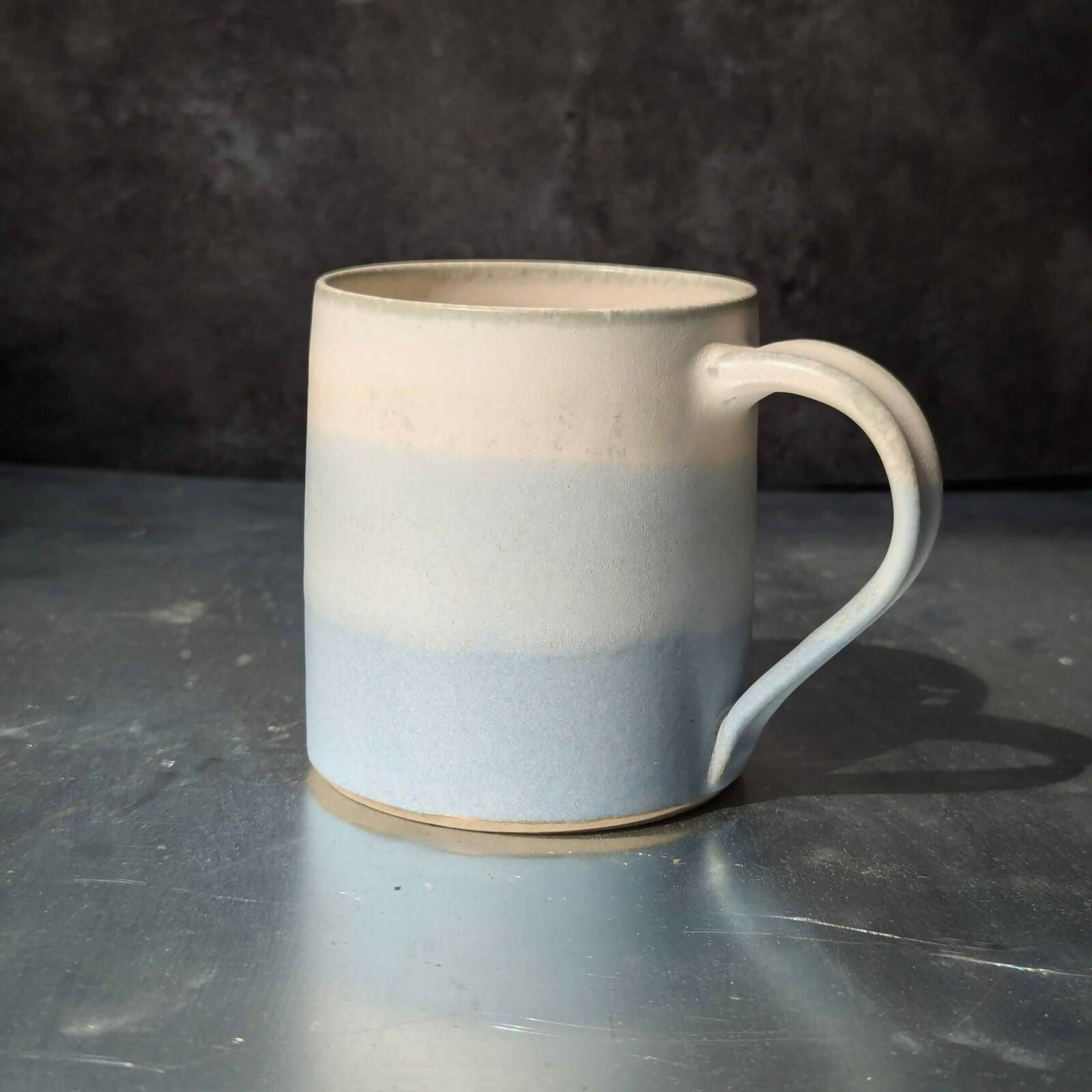 Un mug en grès blanc, tourné et ansé à la main, et émaillé en dégradé de bleu et blanc.